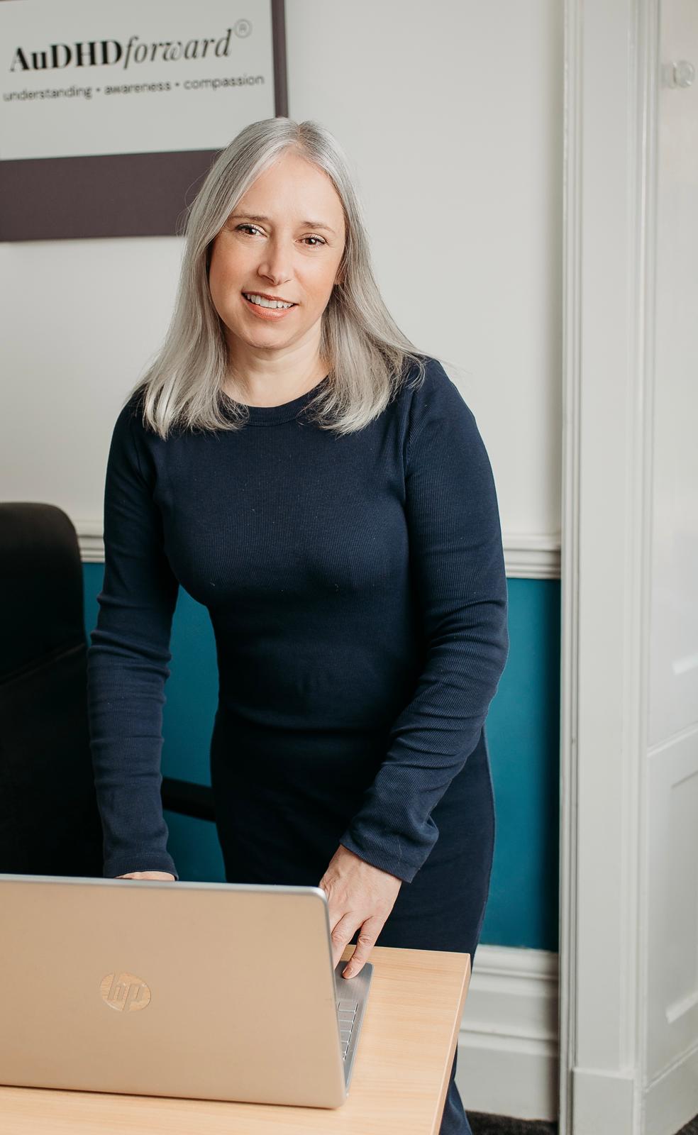 Marie smiling at her desk with a laptop, the AuDHD Forward sign on the wall behind her