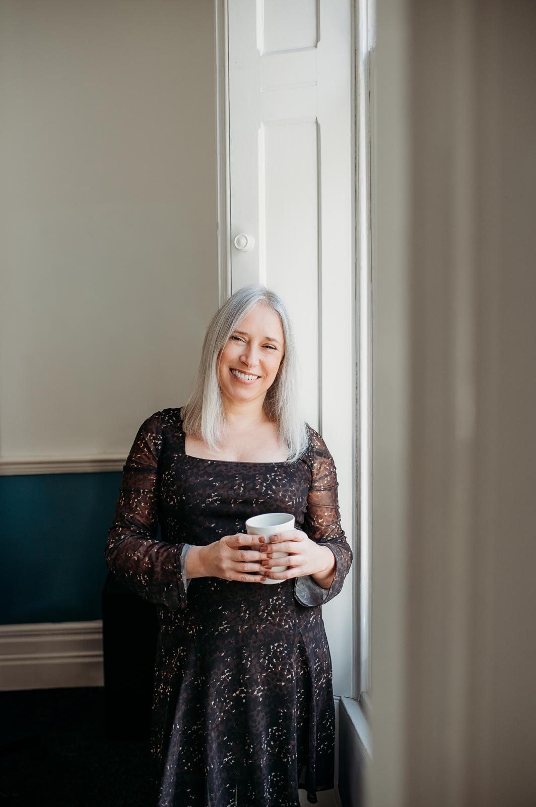 Marie smiling by a window, holding a white mug